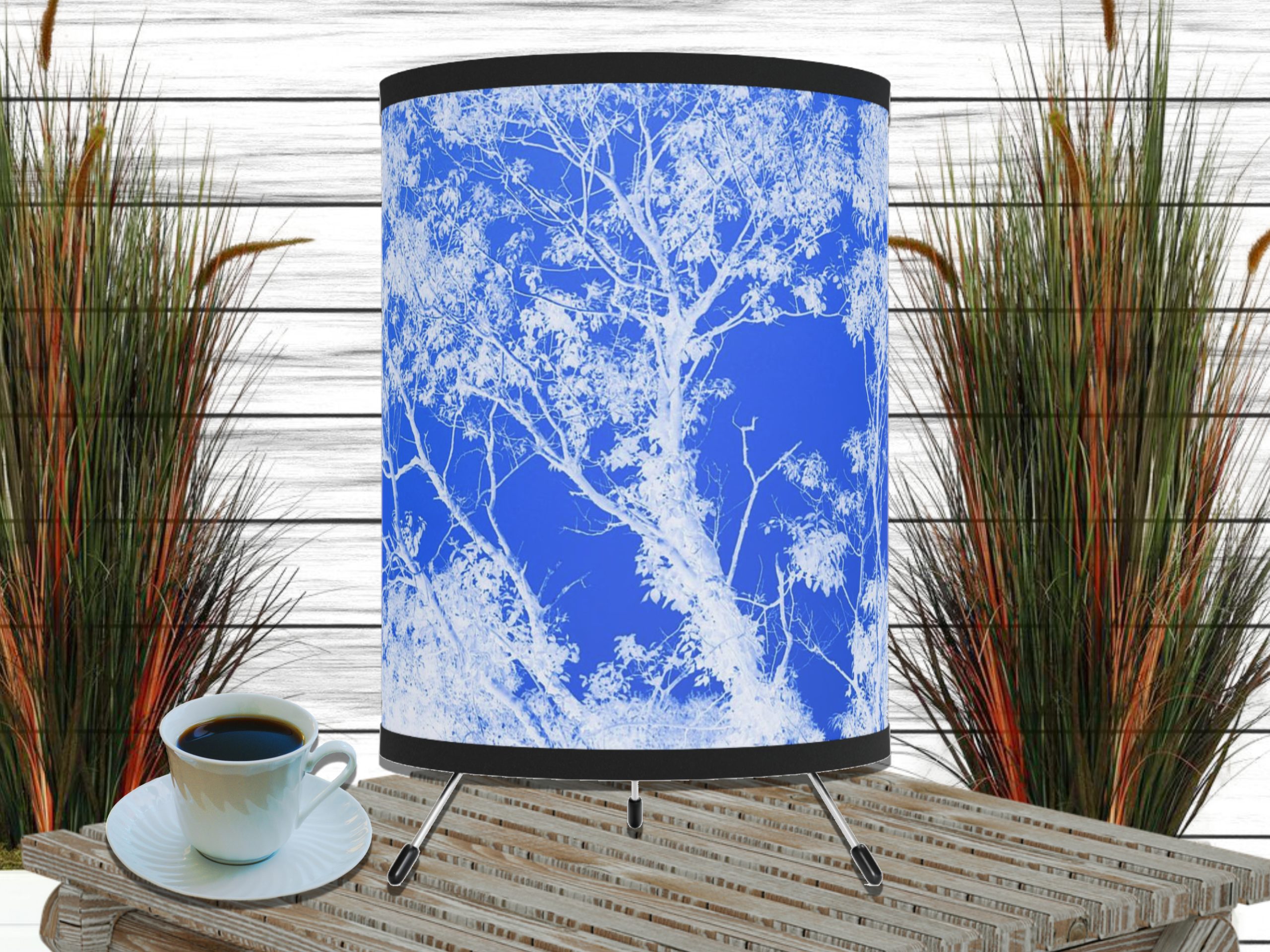 A stylish lamp with a white tree silhouette against a vivid blue background stands on a striped wooden table. Next to it, a cup of black coffee and tall grass plants in soft, natural light.