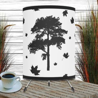 Black tree silhouette with falling leaves on a white lamp, set between tall grasses. A cup of coffee sits on a wooden surface, creating a rustic, cozy ambiance.
