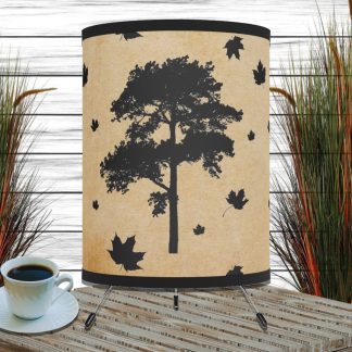 A table lamp with a tree silhouette and falling leaves is on a wooden surface next to a cup of coffee. It’s surrounded by tall, decorative grasses.