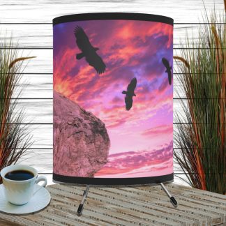 Table lamp with a sunset scene of three silhouetted birds flying over a cliff, set against a vibrant pink and purple sky. Cup of coffee and tall grasses nearby.