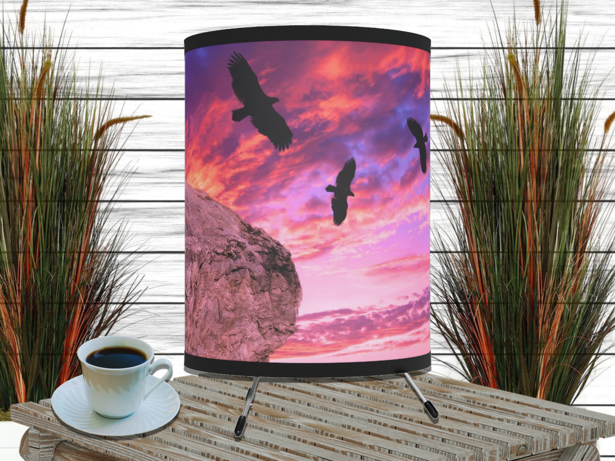 Table lamp with a sunset scene of three silhouetted birds flying over a cliff, set against a vibrant pink and purple sky. Cup of coffee and tall grasses nearby.