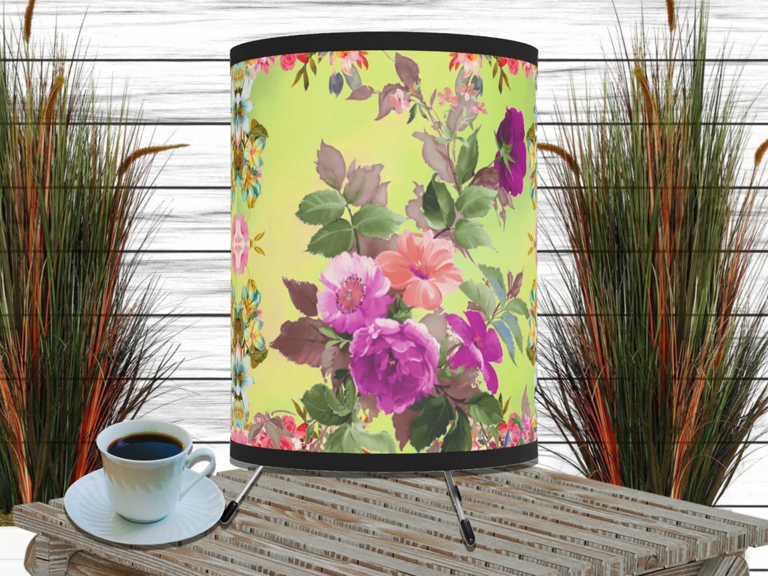 Floral-patterned yellow lampshade on a wooden table with a cup of coffee. Background features tall grass decor against a white-paneled wall.