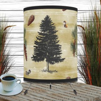 Table lamp with a pine tree and cone design on rustic parchment paper colored lampshade sits on a wooden table beside a cup of black coffee. Tall grass plants flank the scene, creating a cozy ambiance.