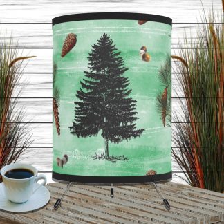 Lampshade with a black tree silhouette and pine cones on a mint green background. It sits on a wicker surface beside a cup of coffee and tall grasses, creating a rustic vibe.