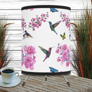 A decorative lampshade with hummingbirds, pink flowers, and butterflies on a white background. Nearby is a coffee cup on a wooden table, with tall grass in the background.