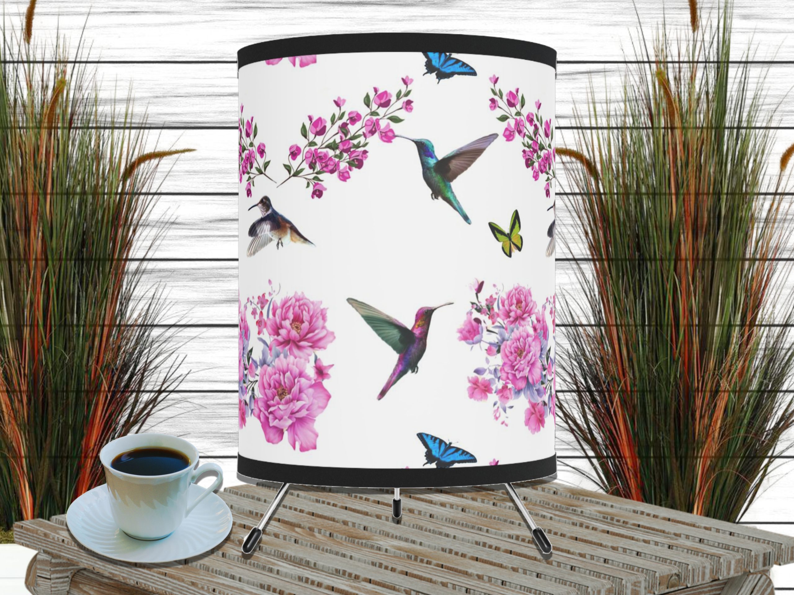 A decorative lampshade with hummingbirds, pink flowers, and butterflies on a white background. Nearby is a coffee cup on a wooden table, with tall grass in the background.