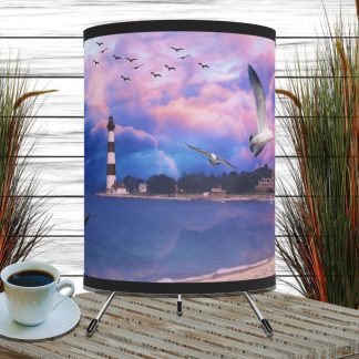 A cylindrical lamp with a lighthouse and seagulls under a colorful sky is placed on a wooden table. A coffee cup sits beside it, flanked by tall grasses.