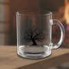 Clear glass mug with intricate black tree design, placed on a wooden surface. A warm, blurred fireplace in the background suggests a cozy atmosphere.