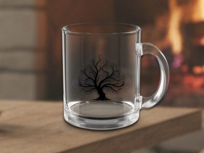 Clear glass mug with intricate black tree design, placed on a wooden surface. A warm, blurred fireplace in the background suggests a cozy atmosphere.
