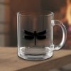 A clear glass mug with a black dragonfly silhouette is placed on a wooden surface. A warm, cozy fireplace creates a blurred, inviting background.
