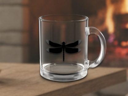 A clear glass mug with a black dragonfly silhouette is placed on a wooden surface. A warm, cozy fireplace creates a blurred, inviting background.