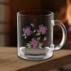 Clear glass mug with pink flowers and green leaves design, placed on a wooden surface near a cozy, blurred fireplace background. Warm and inviting tone.