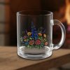 A clear glass mug with a floral design featuring vibrant red, yellow, and blue flowers sits on a wooden table. A cozy fire burns softly in the background.