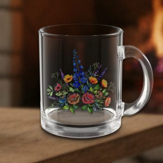 A clear glass mug with a floral design featuring vibrant red, yellow, and blue flowers sits on a wooden table. A cozy fire burns softly in the background.