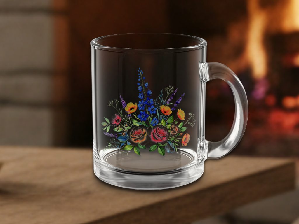 A clear glass mug with a floral design featuring vibrant red, yellow, and blue flowers sits on a wooden table. A cozy fire burns softly in the background.