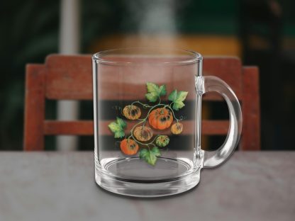 Clear glass mug with painted pumpkins and leaves on a table, faint steam rising, against a warm, blurred chair and background, creating a cozy feel.