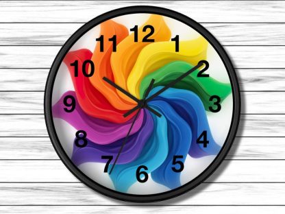A vibrant clock with black numbers and hands features a swirling rainbow design in the center, set against a white wooden background.