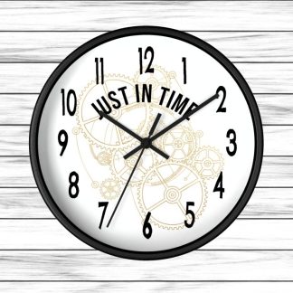 A black wall clock on a white wooden background features black numbers and hands. The center reads "Just In Time" over faint gold clockwork gears.