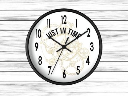 A black wall clock on a white wooden background features black numbers and hands. The center reads "Just In Time" over faint gold clockwork gears.