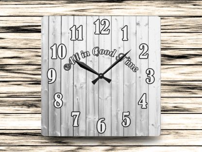 Square wooden clock with large black numbers and the phrase "All in Good Time" across the center. Background features horizontal wooden planks.