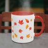 A white mug with a red handle and rim is decorated with orange and yellow autumn leaves. It sits on a table in front of a wooden chair, with steam rising.