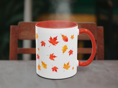 A white mug with a red handle and rim is decorated with orange and yellow autumn leaves. It sits on a table in front of a wooden chair, with steam rising.