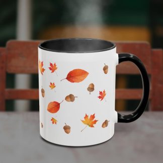 A white mug with black interior, adorned with autumn leaves and acorns, sits on a table. Steam rises from the mug, with a wooden chair in the background.