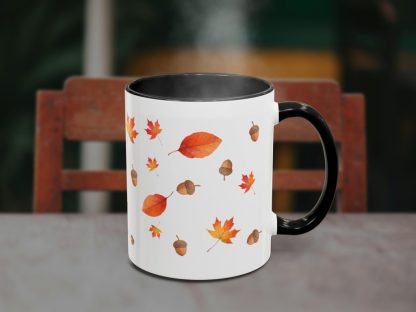 A white mug with black interior, adorned with autumn leaves and acorns, sits on a table. Steam rises from the mug, with a wooden chair in the background.
