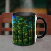 A steaming black mug sits on a table, featuring a vibrant stained-glass forest design with lush green trees and a blue sky, set against a blurred chair.