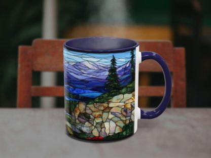 A steaming mug with a stained glass-style design depicting a scenic mountain landscape with trees. The background features a blurred wooden chair.