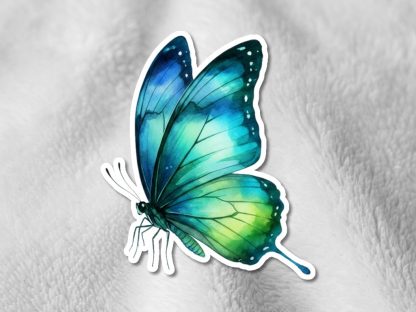 A vibrant, blue and green butterfly sticker with detailed wings is placed on a soft, white textured fabric, creating a serene and delicate contrast.