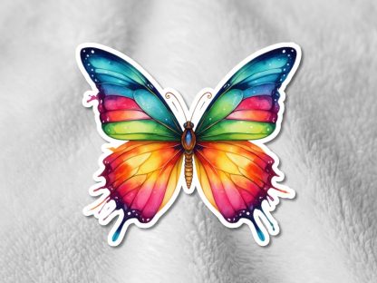 A vibrant butterfly sticker on a soft gray surface. The wings are a gradient of blue, green, pink, and orange, with a dripping paint effect.
