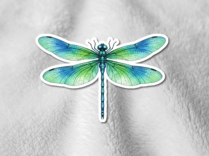 A vibrant dragonfly sticker with intricate green and blue wings is set against a soft, light gray textured fabric background, conveying a serene and natural tone.
