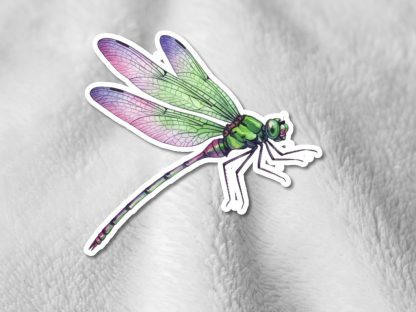 A colorful dragonfly sticker with green and pink wings is placed on a textured, light gray fabric, creating a whimsical and vibrant contrast.