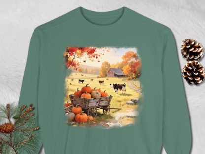 Green sweatshirt with an autumn farm scene. A wagon of pumpkins sits in a field with cows, a barn, and vibrant fall foliage. Cozy and rustic theme.