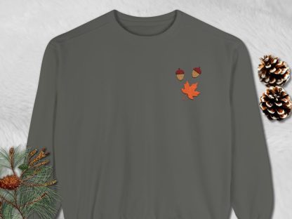 Dark gray sweatshirt on textured gray background. Decorative fall-themed patches: two acorns and a maple leaf in red and orange tones add a seasonal touch.