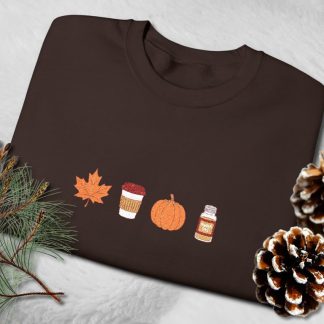 Brown sweatshirt featuring autumn-themed embroidery: a maple leaf, a to-go coffee cup, a pumpkin, and a spice jar. Cozy, seasonal design.