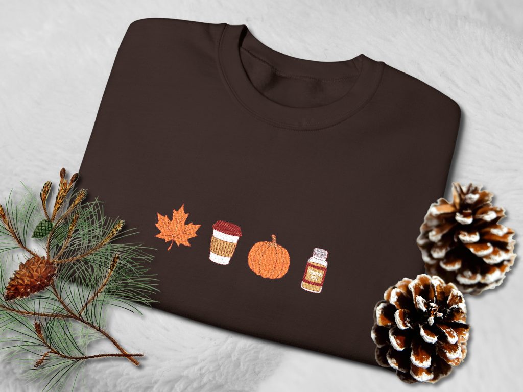 Brown sweatshirt featuring autumn-themed embroidery: a maple leaf, a to-go coffee cup, a pumpkin, and a spice jar. Cozy, seasonal design.