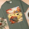 T-shirt featuring an autumn farm scene with pumpkins in a cart, cows grazing, and a barn. Nearby, a coffee cup and pine branch rest on a wooden surface.