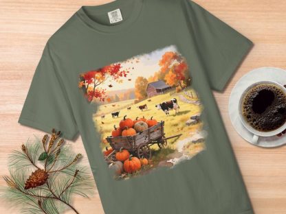 T-shirt featuring an autumn farm scene with pumpkins in a cart, cows grazing, and a barn. Nearby, a coffee cup and pine branch rest on a wooden surface.