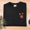 Black t-shirt with embroidered acorns and an orange maple leaf, placed on a wooden table. Nearby are pine branches with a cone and a cup of coffee.