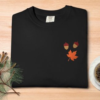 Black t-shirt with embroidered acorns and an orange maple leaf, placed on a wooden table. Nearby are pine branches with a cone and a cup of coffee.
