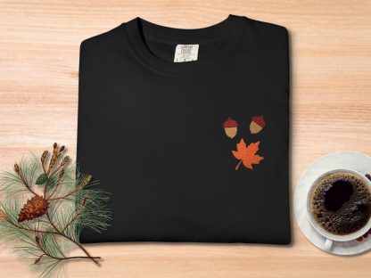 Black t-shirt with embroidered acorns and an orange maple leaf, placed on a wooden table. Nearby are pine branches with a cone and a cup of coffee.