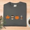 Gray T-shirt with autumn icons: orange leaf, coffee cup, pumpkin, and pumpkin spice jar. Set on a wood table with pine branches and coffee cup. Cozy fall vibe.