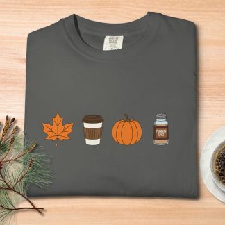 Gray T-shirt with autumn icons: orange leaf, coffee cup, pumpkin, and pumpkin spice jar. Set on a wood table with pine branches and coffee cup. Cozy fall vibe.