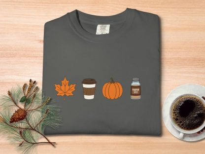 Gray T-shirt with autumn icons: orange leaf, coffee cup, pumpkin, and pumpkin spice jar. Set on a wood table with pine branches and coffee cup. Cozy fall vibe.