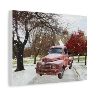 A vintage red truck is parked on a snowy path between trees, with a backdrop of a brick building. Snow gently falls, creating a serene winter scene. Canvas wall art.