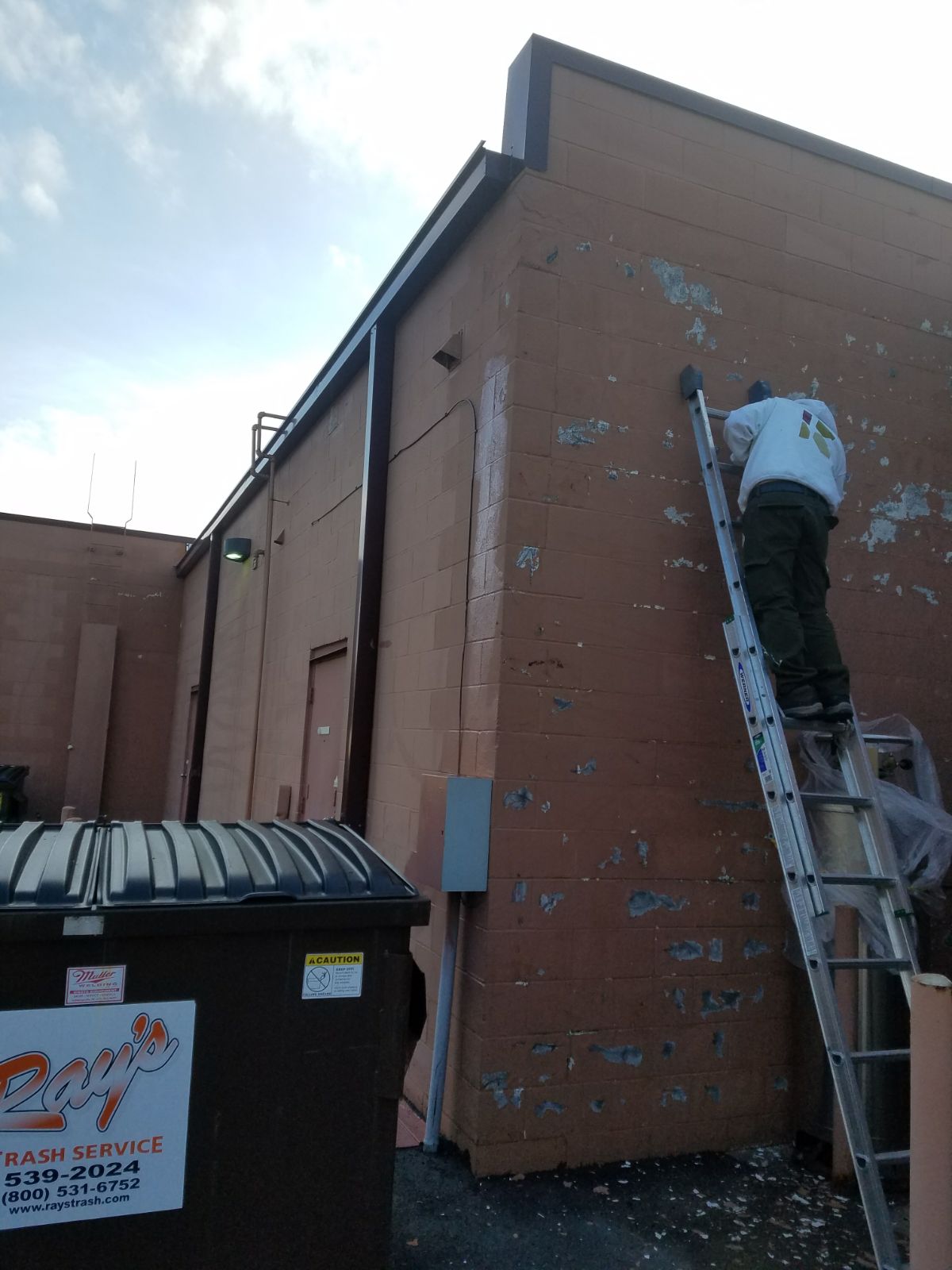 Professional painter using a ladder to prepare and repaint a commercial building's exterior wall.