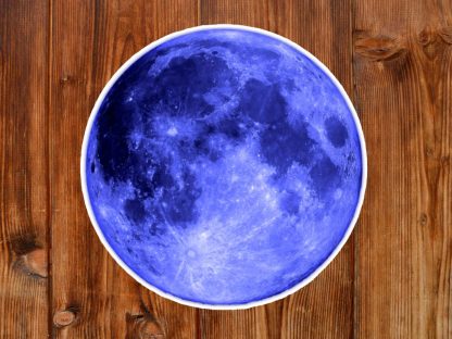 Gothic sticker of a round vivid blue moon design sitting on a wooden table, combining a celestial theme with rustic texture, creating a mystical, earthy ambiance.