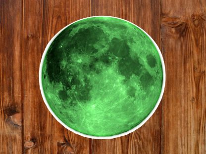 Gothic round sticker of a green moon placed on a rustic wooden surface. The vibrant green tones contrast with the warm, textured wood, creating a mystical atmosphere.